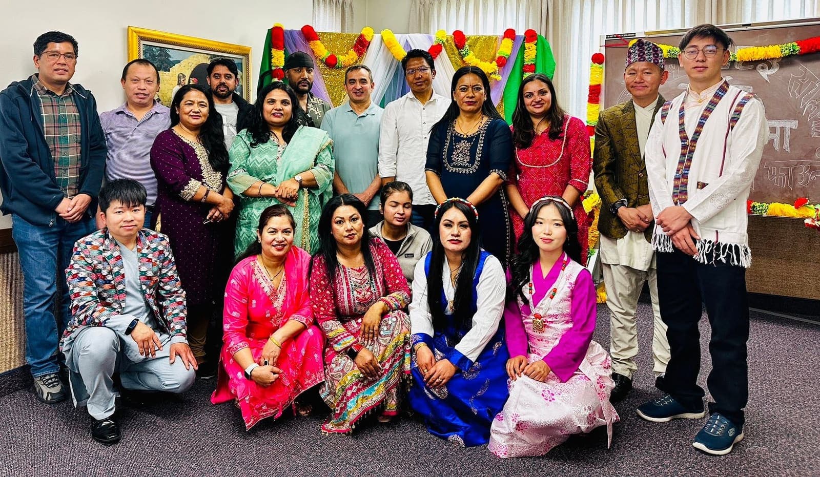 Around twenty members of the community together in a decorated hall, with Nepali writing on the board behind them