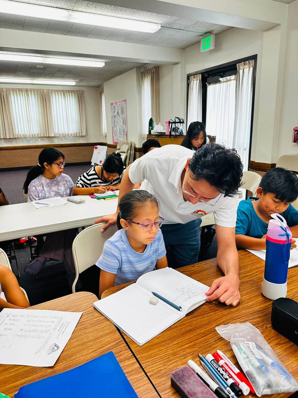 A volunteer teacher in a Community Rise Foundation shirt leaning over a table to help a girl with her Nepali handwriting