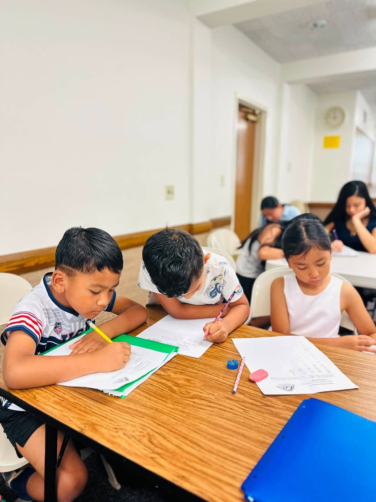 Three children copying Devanagari letters onto worksheets at a classroom table